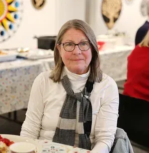 Woman seated at a decorated table, wearing glasses and a gray scarf in a bright dining room.