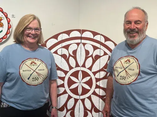 Two smiling people in light blue shirts stand before a large red-and-white circular pattern.