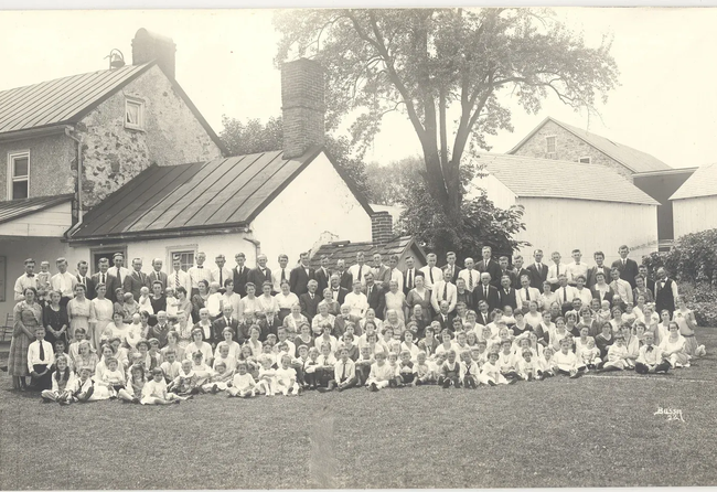 Large group gathered outdoors in front of stone buildings and trees, posing for a historic group photo.