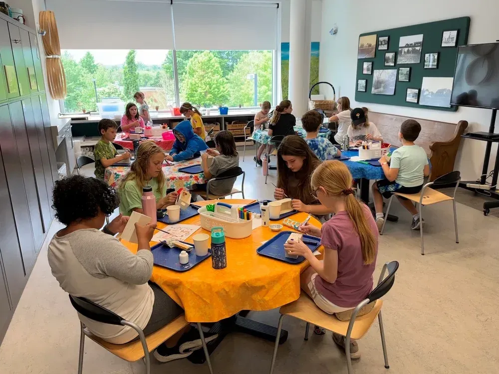 Students working at orange-covered tables in a classroom, with art supplies and a teacher nearby.