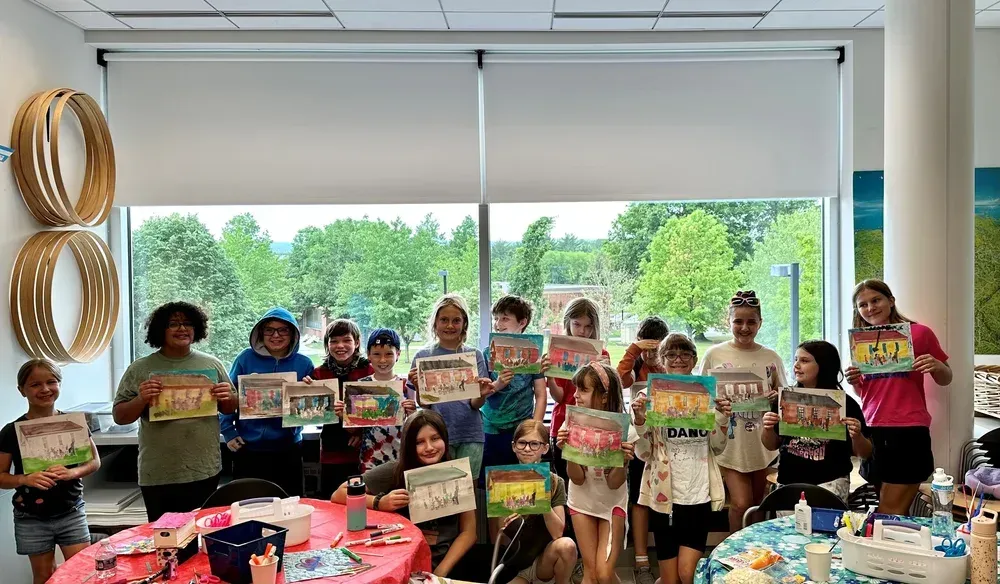 Children holding colorful signs in a bright classroom or event space, standing behind tables with snacks.