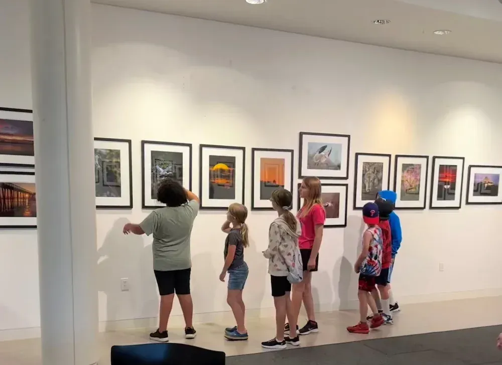 Children viewing framed artworks in a gallery, with one person pointing at a picture.