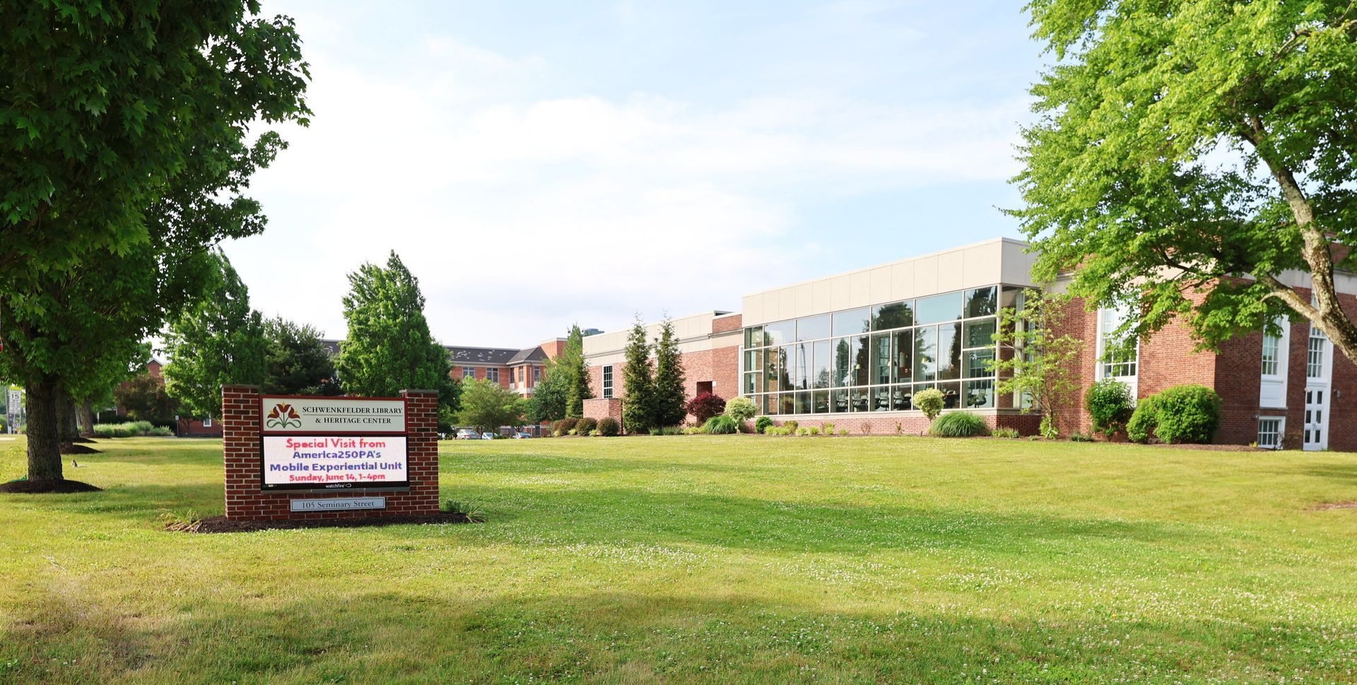 Campus lawn with trees, brick buildings, and a sign near a modern glass-fronted building.