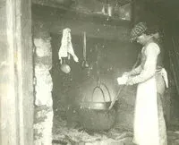 Person in a kitchen stirring a hanging pot over a fire, with clothes hanging nearby.