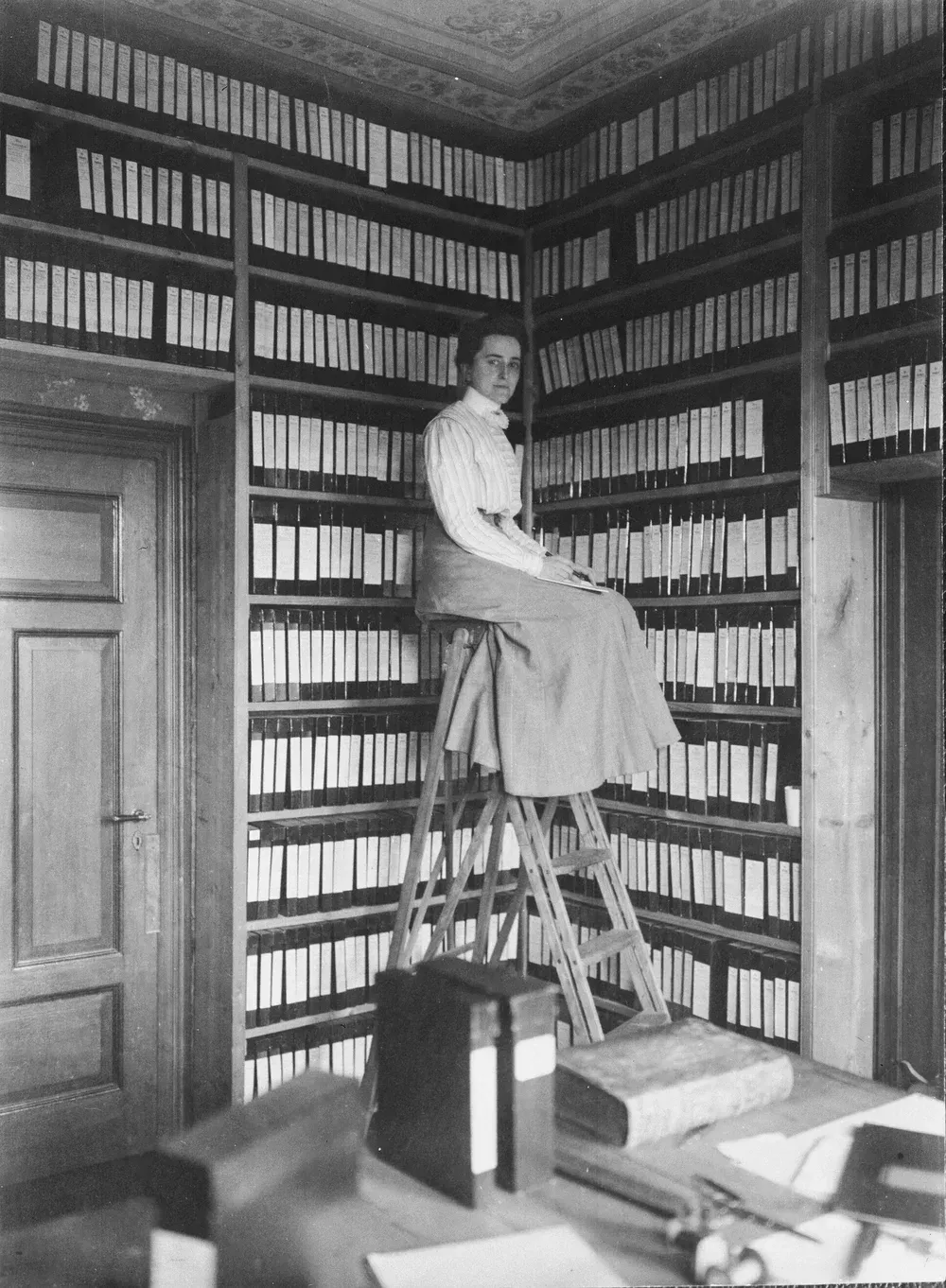 Person perched on a ladder in a book-lined room, reaching among tall shelves