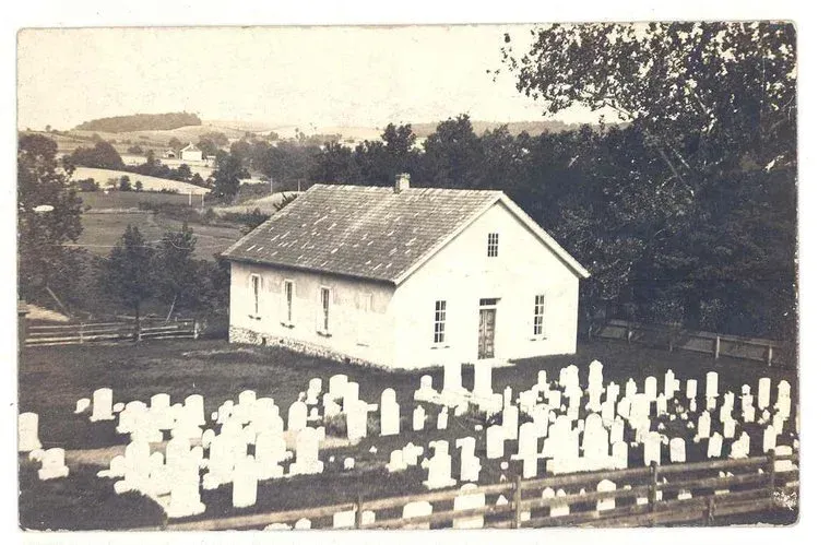 Small white chapel beside a graveyard, with rows of white headstones in a rural landscape.