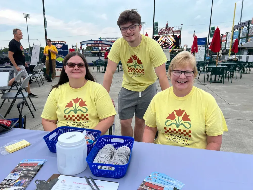 Three volunteers in yellow T-shirts at a table outdoors, with donation baskets and a building in the background.