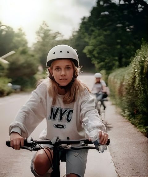 Child wearing a white helmet rides a bike on a tree-lined path with another cyclist behind.