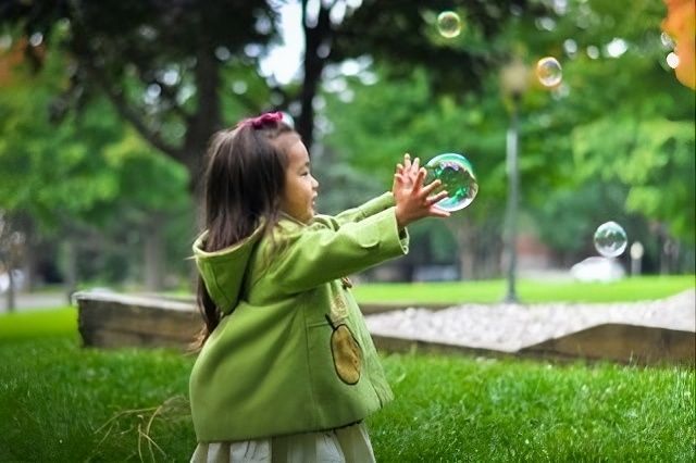 Child in a green sweater reaching for soap bubbles in a grassy park