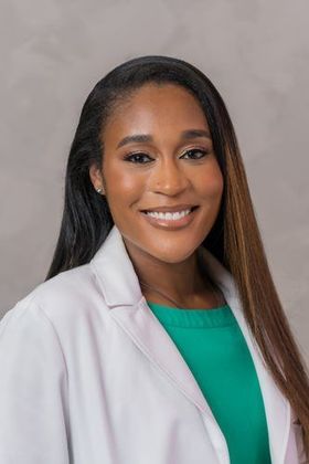 Smiling professional in a white coat and green top against a neutral gray backdrop