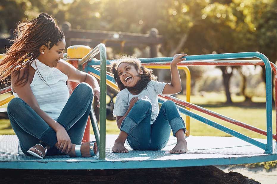 A woman and a child laugh while sitting on a colorful merry-go-round at a playground on a sunny day.