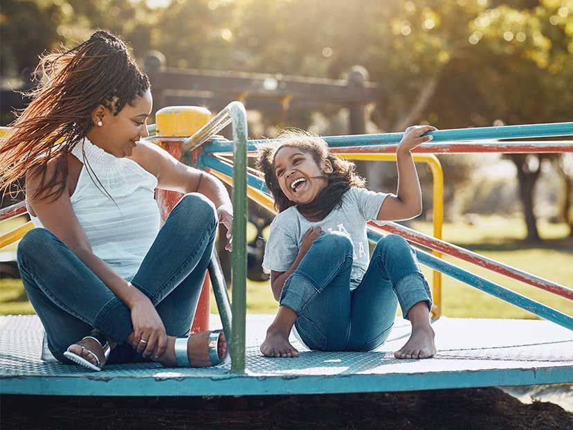 A woman and a child smiling and playing on a playground merry-go-round in sunlight
