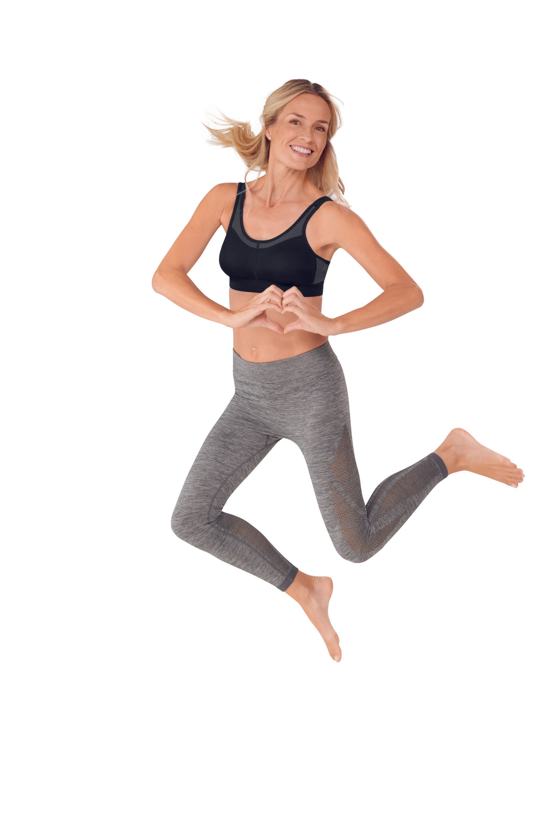 Smiling woman in black sports top and gray leggings jumping against a white background