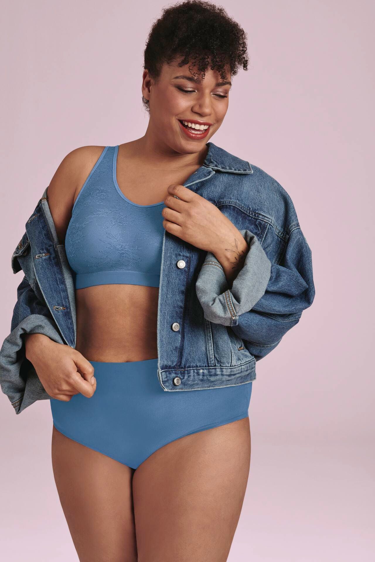 Person in blue lingerie and denim jacket against a pink background, looking down and smiling.