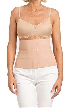 Woman wearing a beige torso support brace over a bra and white pants, standing against a white background