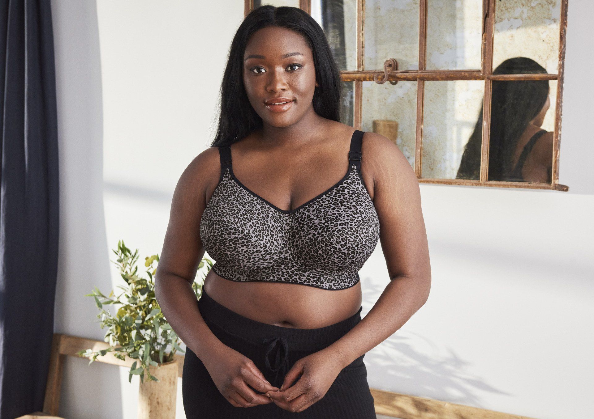 Woman standing indoors by a window, wearing a patterned crop top and black pants, with a plant nearby.