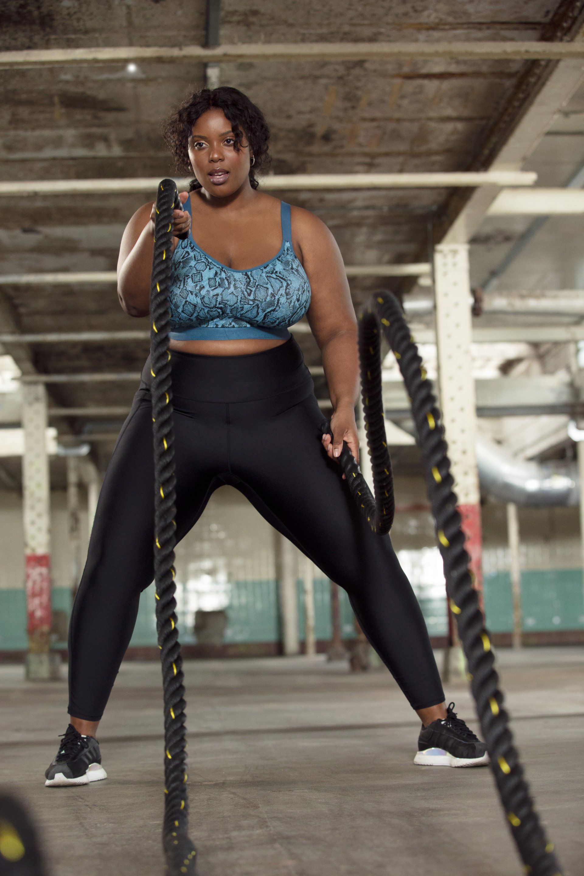 Woman exercising with battle ropes in a parking garage, wearing black leggings and a blue sports bra.