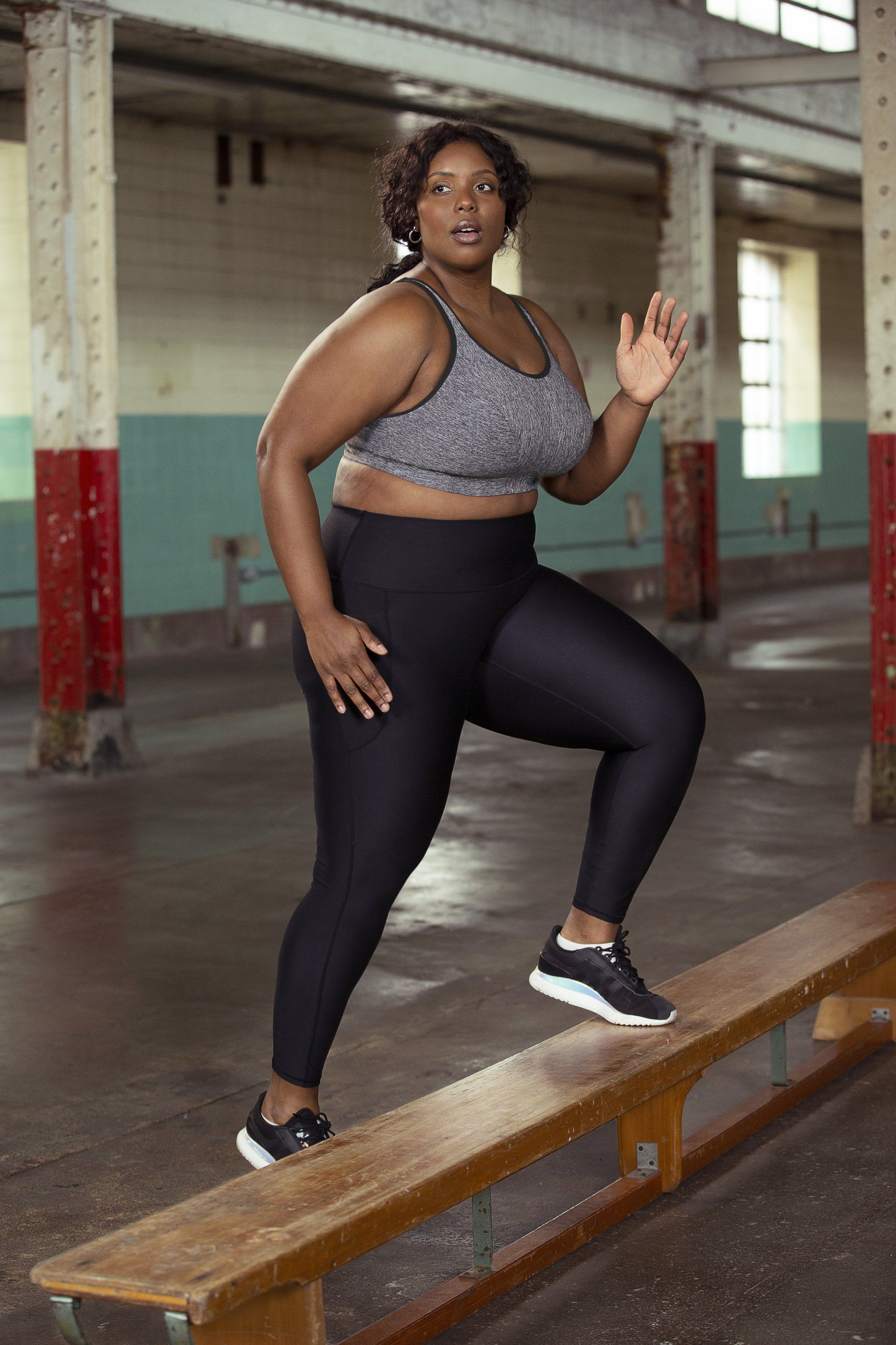 Person poses on a bench in an industrial-looking room, wearing black leggings and sneakers.