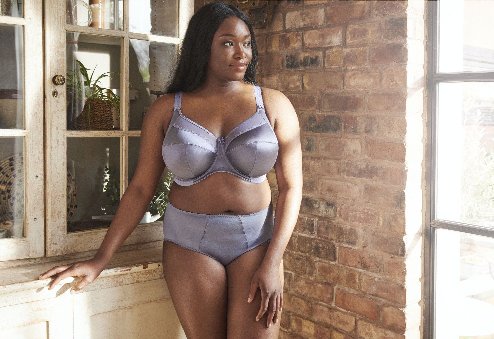 Woman in lavender lingerie standing by a window in a brick-walled room