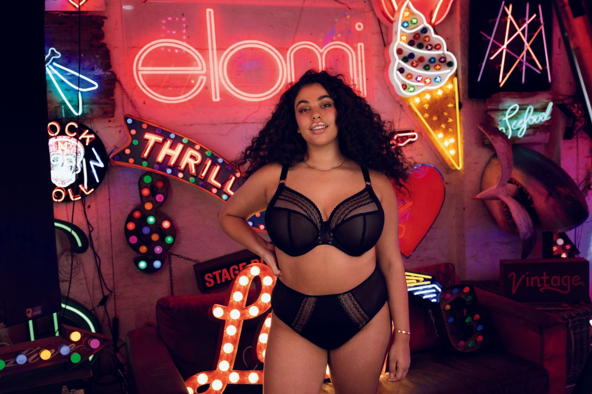 Woman in black lingerie posing in a neon-lit room with retro signs and colorful lights.