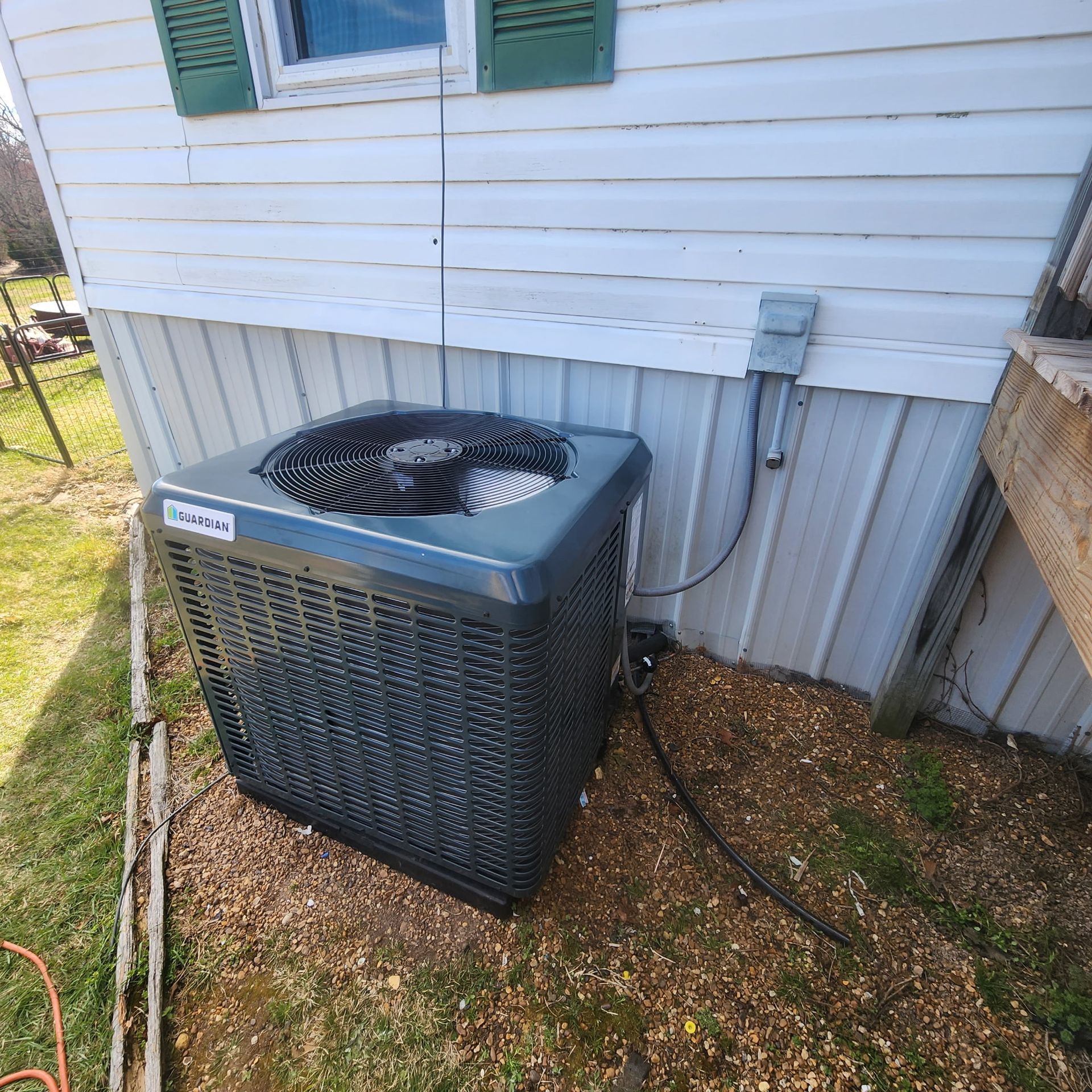 An air conditioning unit next to a white house with green shutters and brown ground cover.