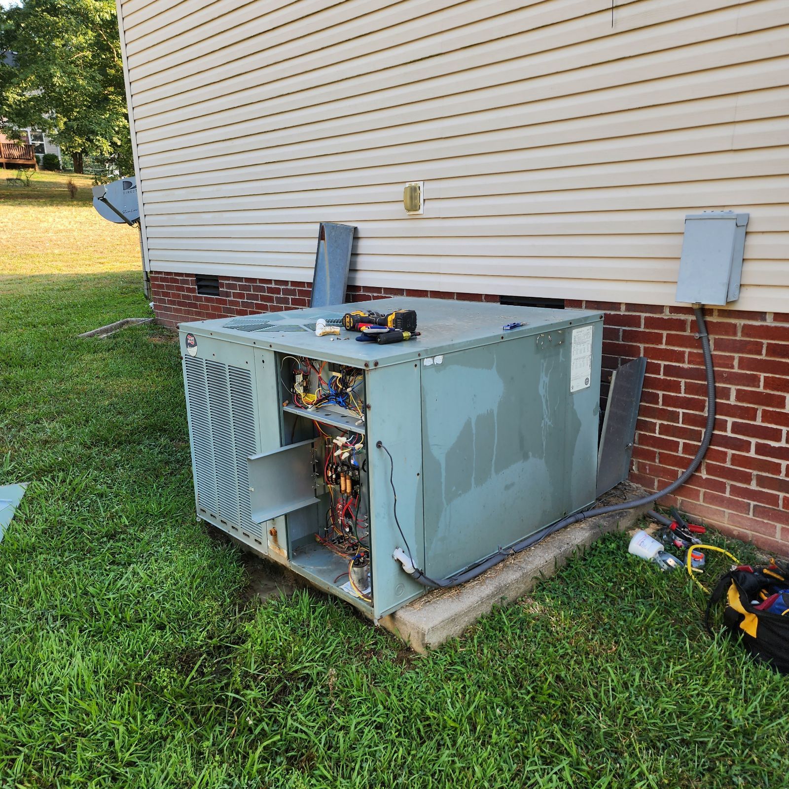 Open air conditioner unit next to a house with exposed wiring and tools on top.