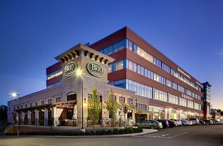 Modern office building at dusk with brick and glass facade, lit windows, and a parking lot outside