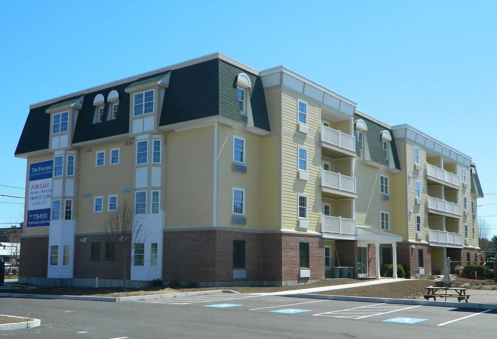 Three-story beige apartment building with green roof trim and balconies on a sunny day.