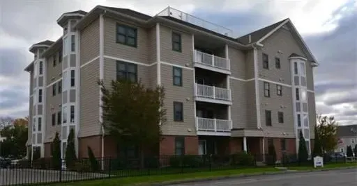 Four-story beige apartment building with balconies and a grassy lawn under cloudy skies
