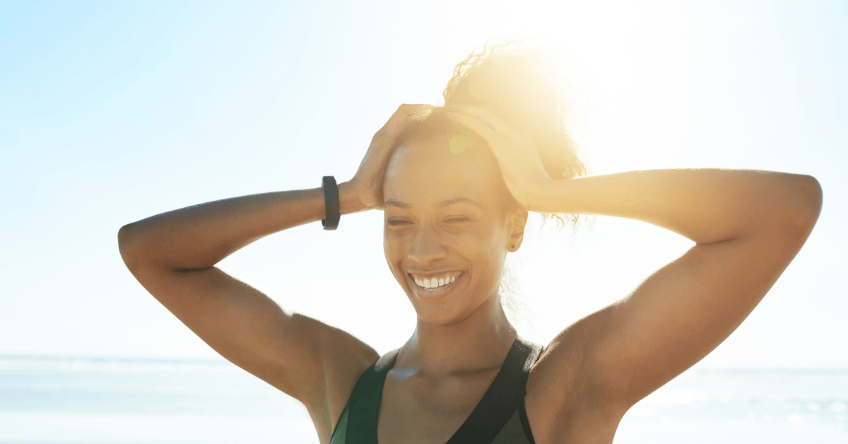 A woman is smiling with her hands behind her head on the beach.