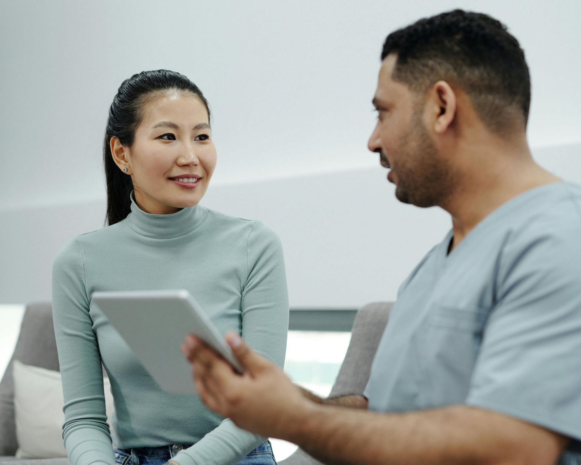A person in scrubs shows a tablet to a seated person in a waiting room, both looking at each other.