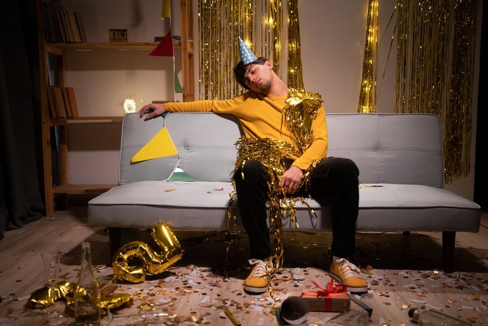 A man is sitting on a couch in a messy room after a party.