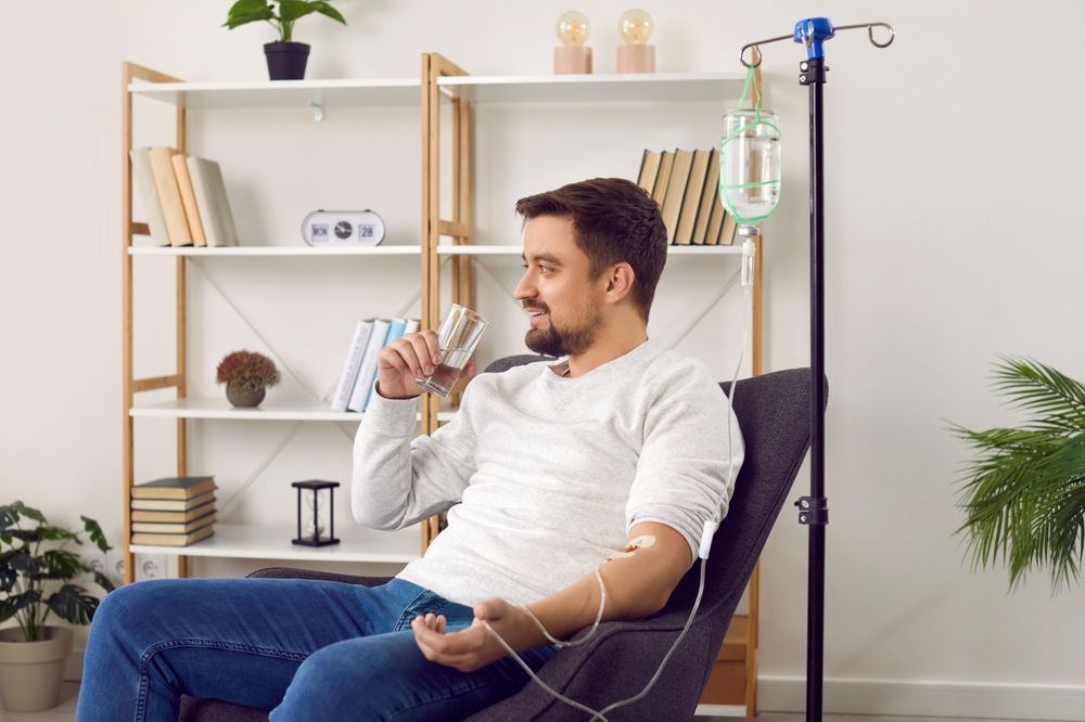 A man is sitting in a chair with an iv in his arm and drinking a glass of water.