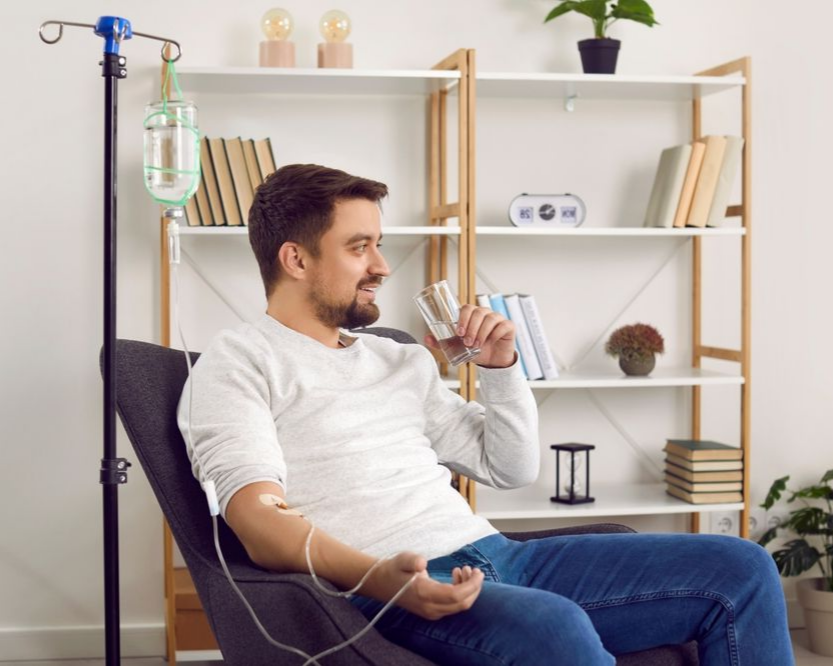 Man receiving IV treatment, sitting in chair, smiling. IV bag on stand, modern room.
