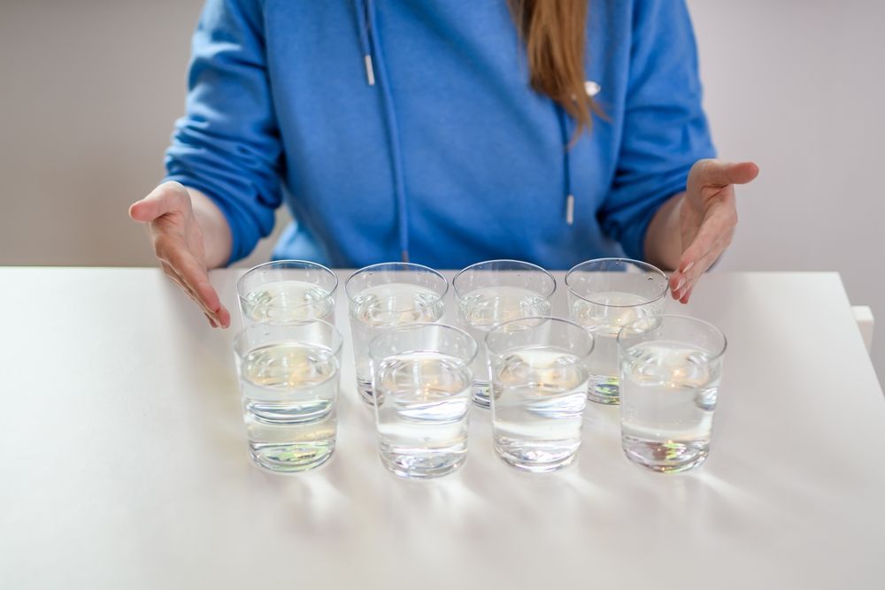 A woman is sitting at a table with six glasses of water.