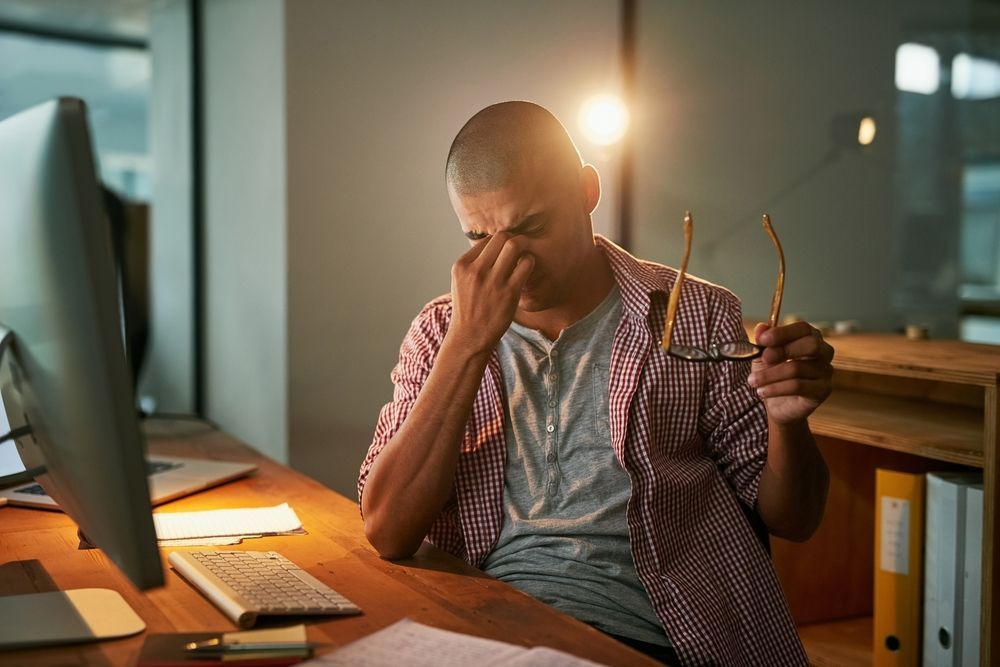 A man is sitting at a desk with a computer and rubbing his eyes.
