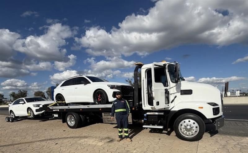 Tow truck loading a white car on a flatbed, with another white car parked nearby under a cloudy sky.