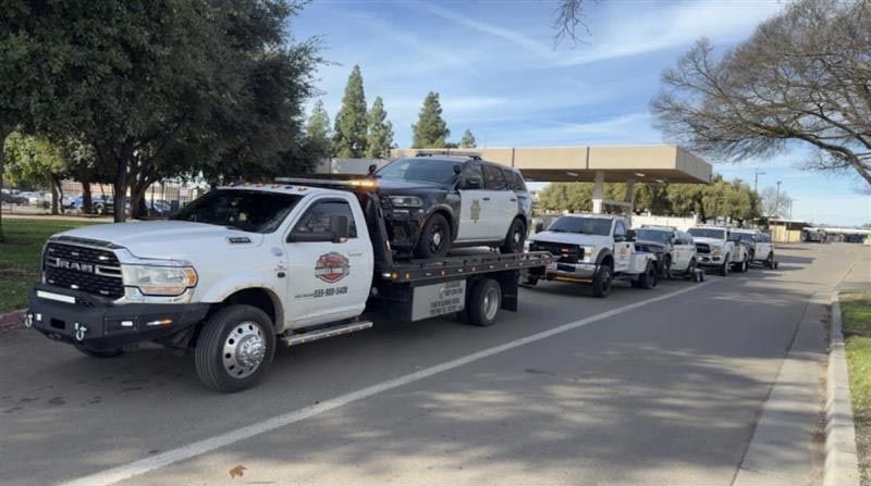Line of white tow trucks parked along a street beside a building and trees