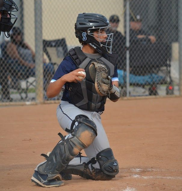 Youth baseball catcher crouched behind home plate, in black gear and gray pants, ready to catch a pitch.