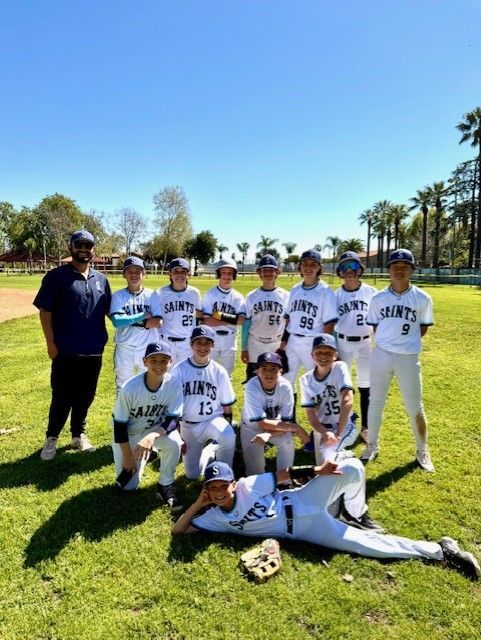 Baseball team posing on a sunny field, wearing white uniforms, with a coach and a trophy in front.