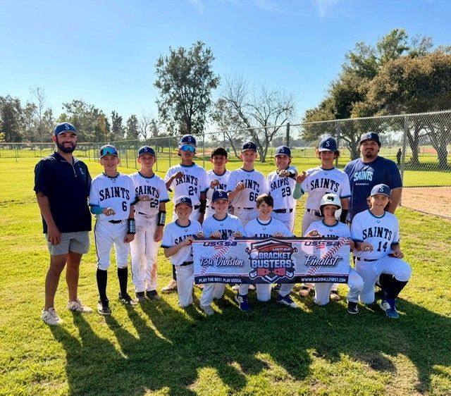 Youth baseball team in white and blue uniforms posing with coaches on a sunny field, holding a championship banner