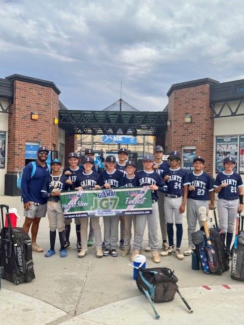 Youth baseball team posing with coach at a stadium entrance, holding a banner and gear bags nearby.