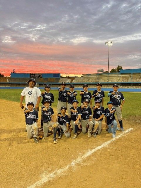 Youth baseball team posing on a dirt field at sunset, with pink-orange sky and stadium bleachers behind them.