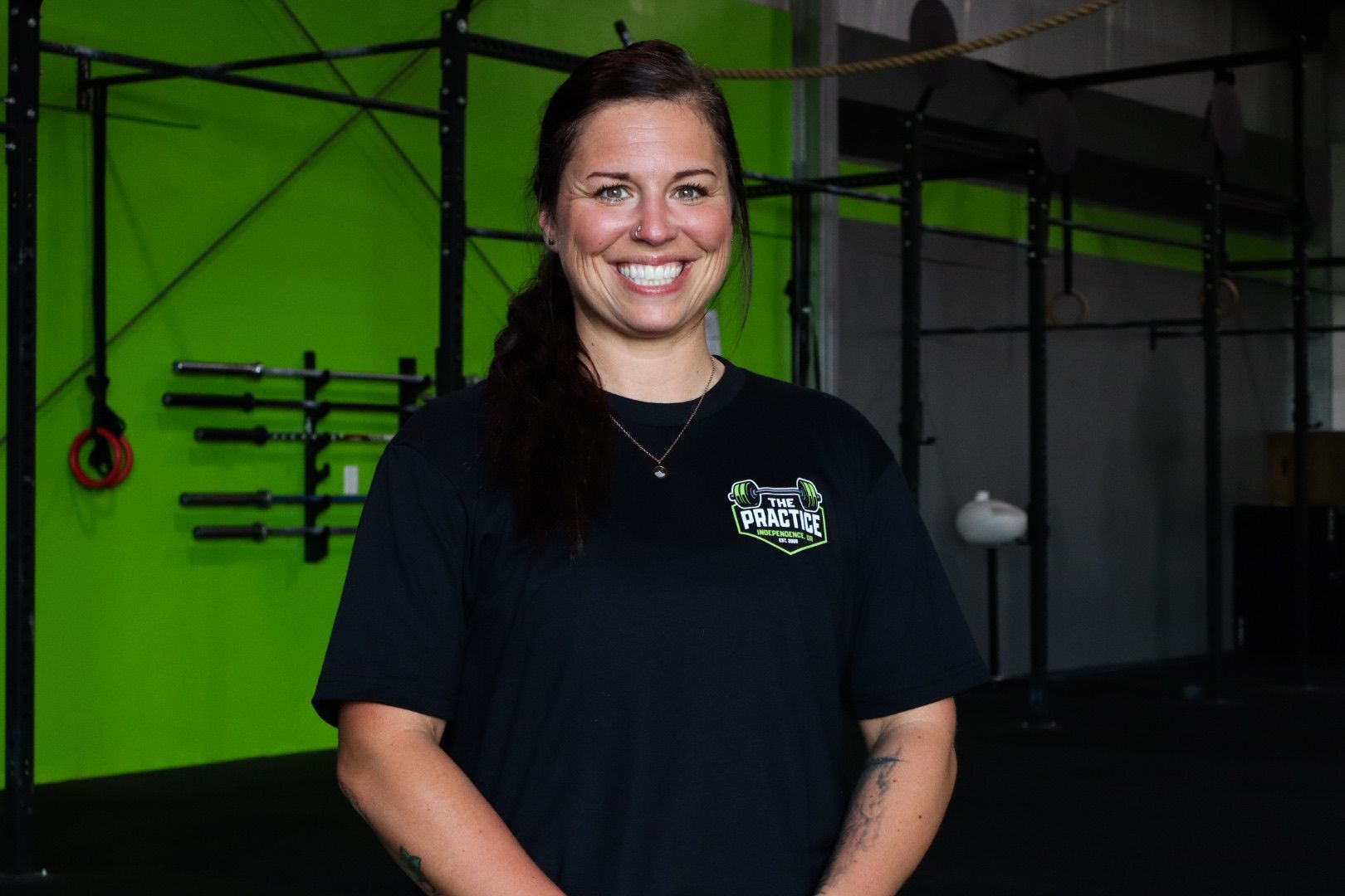 Smiling person in a black shirt standing in a gym with green walls and workout equipment.