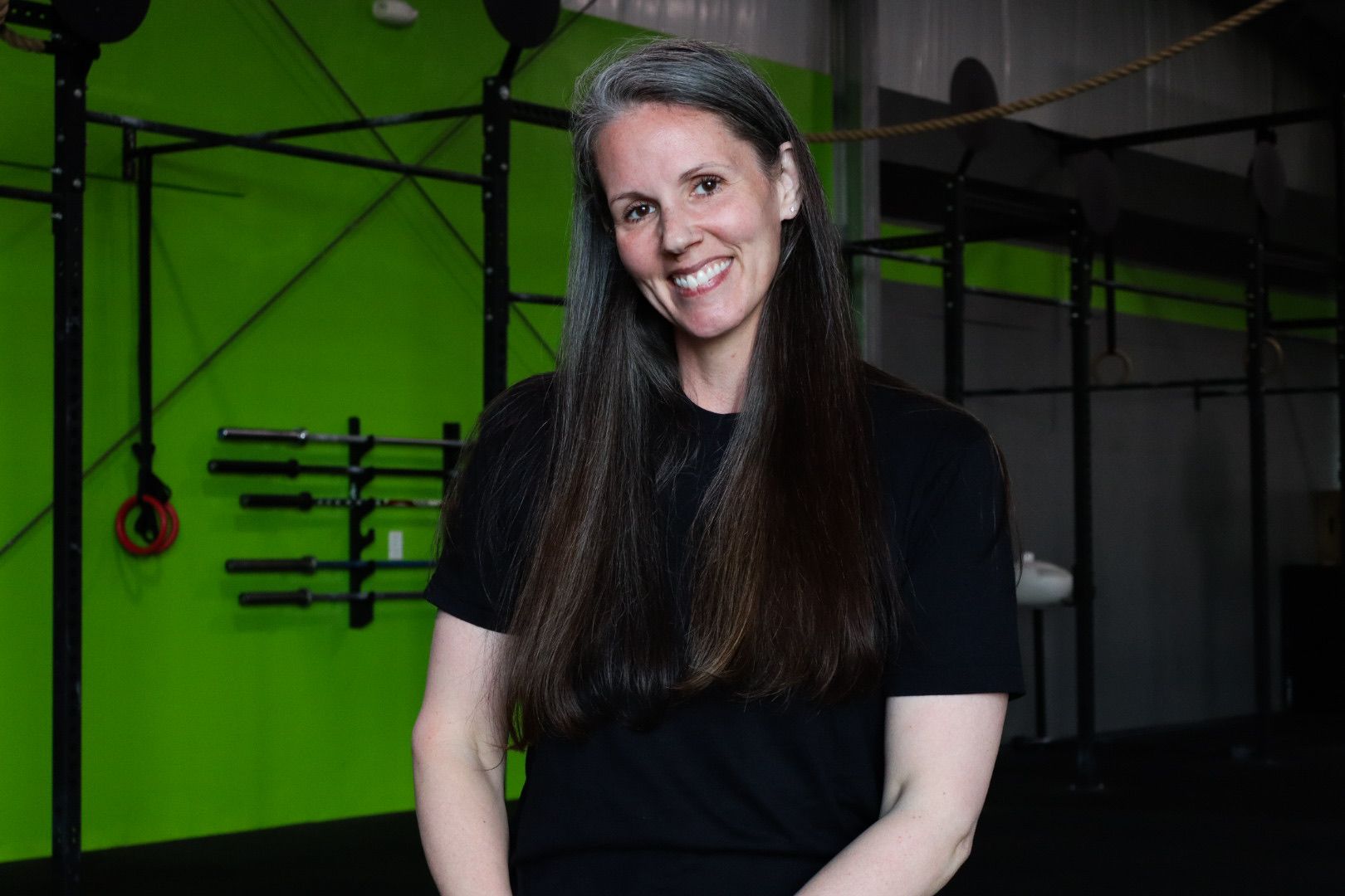 Woman with long, gray hair smiles, wearing a gray sweatshirt with design, sitting on a wooden box in a gym.
