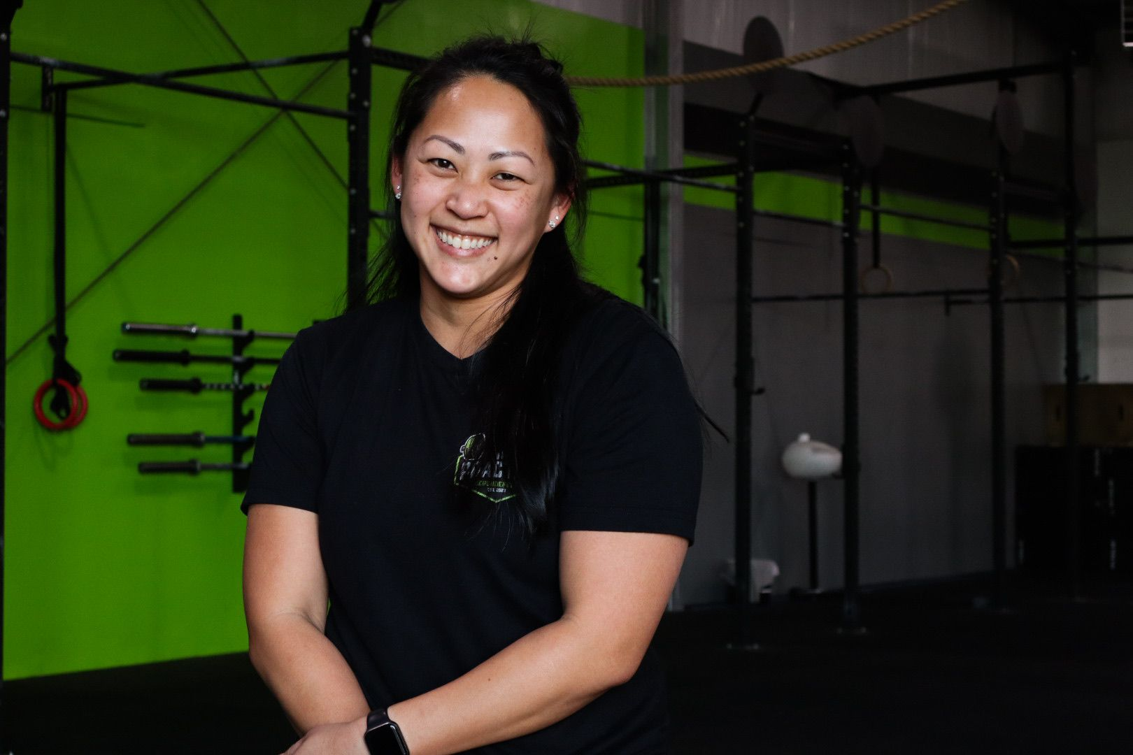 Woman in black shirt smiles, leaning on weight bar with plates in a gym. Yellow wall in background.