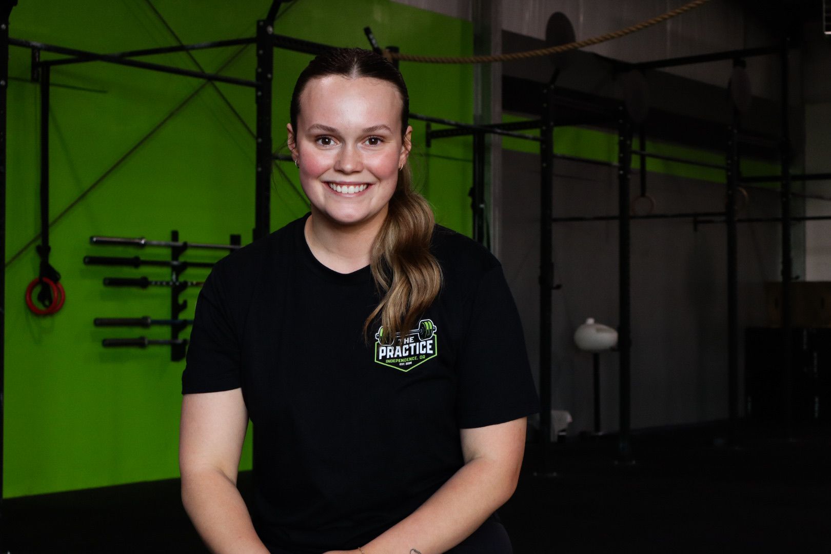 Smiling person seated in a gym with a bright green wall and pull-up bars behind them