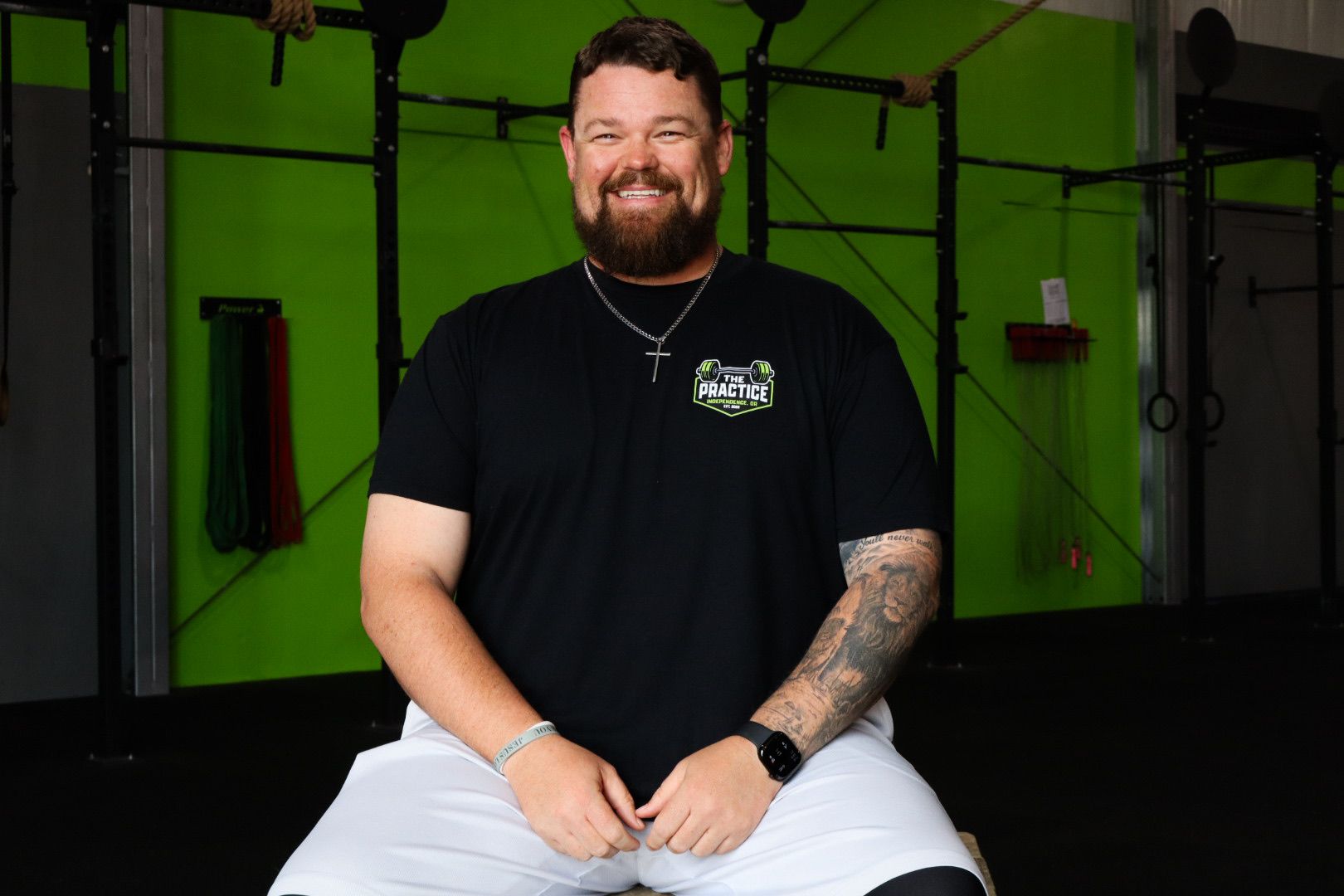 Smiling man seated in a gym, wearing a black T-shirt and white shorts, with workout equipment behind him