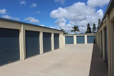 Rows of beige storage units with dark blue roller doors on a concrete surface under a partly cloudy sky.