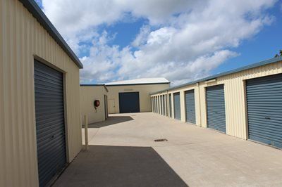 Rows of beige storage units with gray roll-up doors under a partly cloudy sky. A concrete path leads between the units.